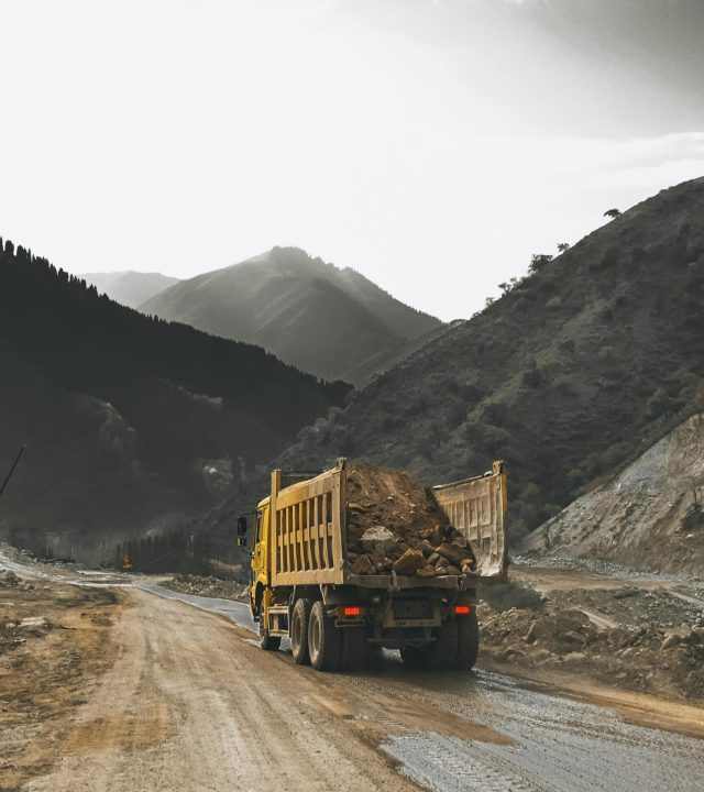 A yellow dump truck carrying dirt on a winding unpaved road through mountainous terrain.